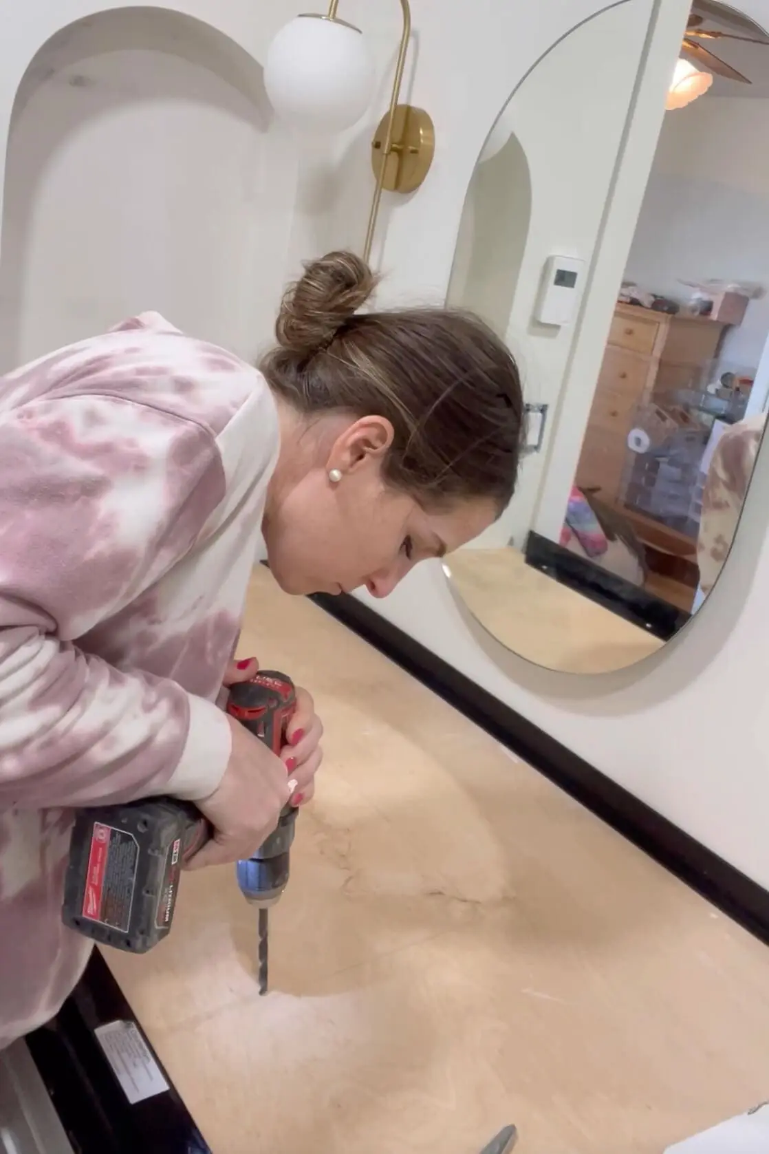 A woman drilling a hole into a wooden countertop base using a red power drill. The workspace includes a black plastic edge frame, a gold wall sconce, and an oval mirror on the wall, with household items visible in the background.
